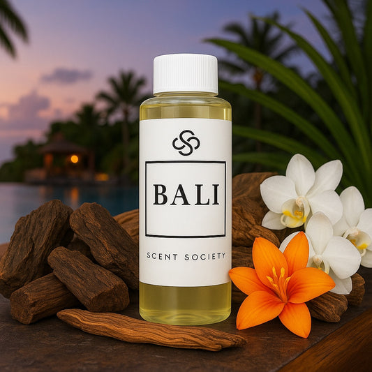 Scent Society BALI premium diffuser fragrance oil in a clear 100ml plastic bottle with white label and ap, placed on a stone surface beside vibrant orange flowers, with tropical resort pool and palm trees at sunset in the background.