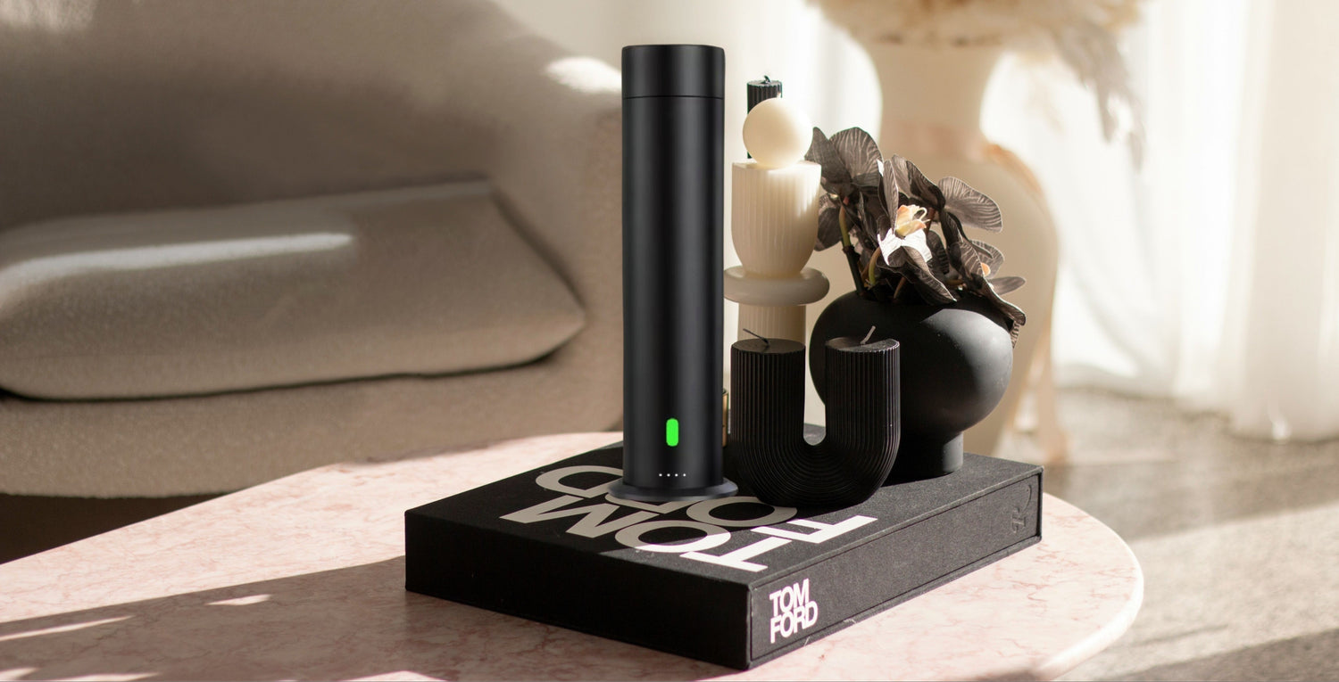 Store hero image banner. Black electronic device on a marble coffee table and wooden book stand with 'Tom Ford' branding in a minimalistic setting. White pampas in a beige jumbo vase in background next to plush beige sitting chair. White curtains with sunlight peeking through.