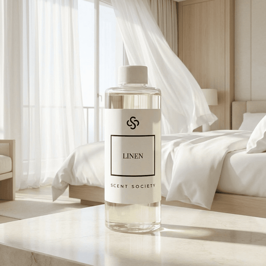 Bottle of Scent Society Linen diffuser oil on a nightstand in a bedroom with white curtains flowing in the background.