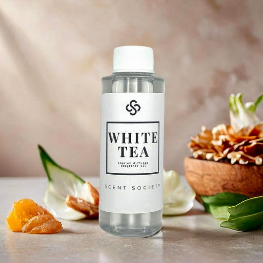 Scent Society WHITE TEA premium diffuser fragrance oil in a clear 100ml plastic bottle with white label and cap, displayed on a stone surface with dried orange peel, white petals, green leaves, and a wooden bowl filled with dried tea leaves in the background. Scent inspired by Westin Hotel.