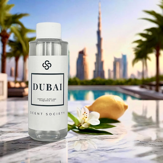 Scent Society DUBAI premium diffuser fragrance oil in a clear 100ml plastic bottle with white label and cap, placed on a white marble surface next to a lemon and white flower with the Dubai skyline and Burj Khalifa in the background. Scent inspired by Delano hotel.