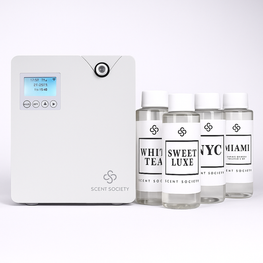 White square diffuser machine with Scent Society logo on bottom right corner. Four 100ml bottles of diffuser oils with white labels and white caps with fragrance names White Tea, Sweet Luxe, NYC, Miami