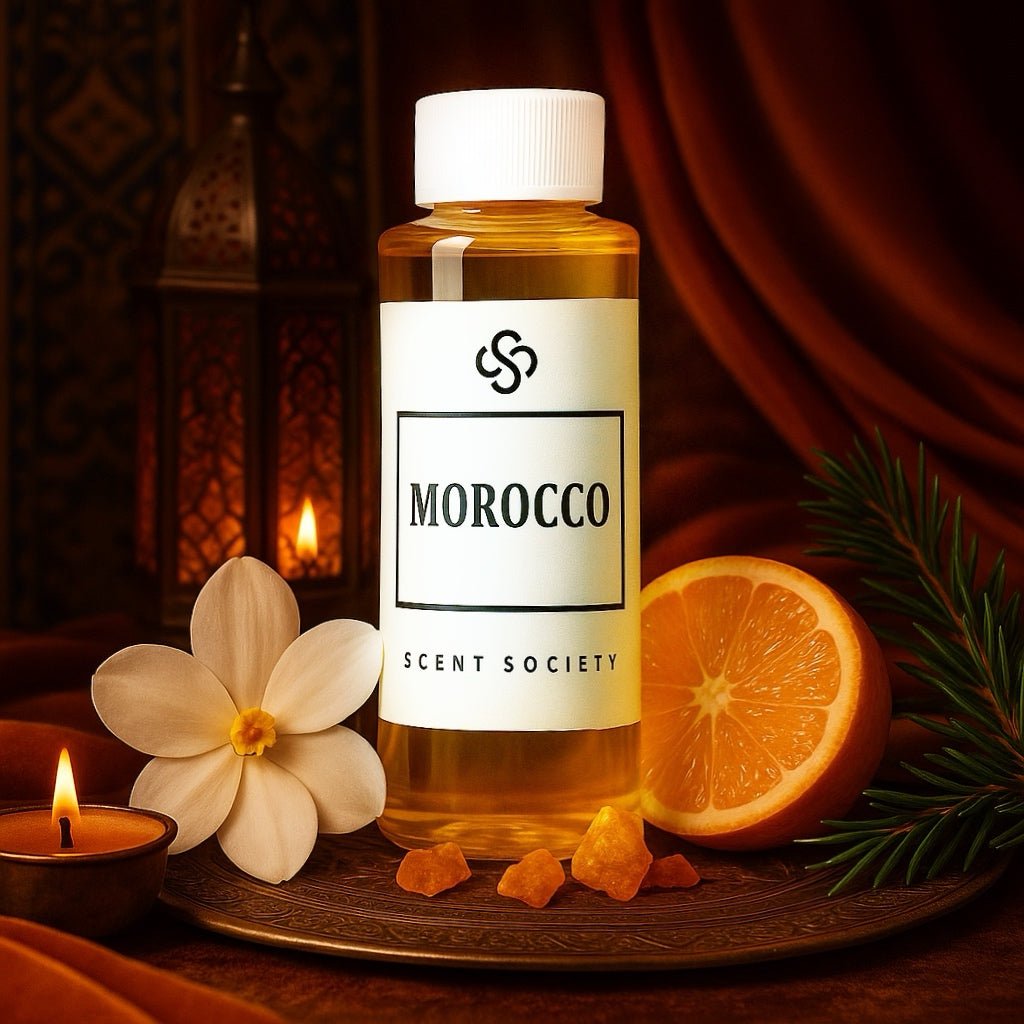 Scent Society Morocco diffuser oil in a 100ml clear bottle with white label and black text, placed on an ornate brass tray surrounded by orange slices, pine sprigs, gardenia flower, amber resin, and a lit candle. The backdrop features rich red drapery and a Moroccan-style lantern, evoking a warm, exotic ambiance.