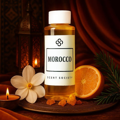 Scent Society Morocco diffuser oil in a 100ml clear bottle with white label and black text, placed on an ornate brass tray surrounded by orange slices, pine sprigs, gardenia flower, amber resin, and a lit candle. The backdrop features rich red drapery and a Moroccan-style lantern, evoking a warm, exotic ambiance.