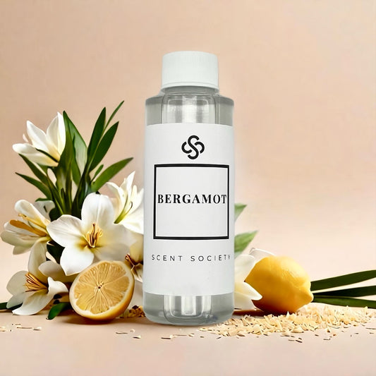 Scent Society BERGAMOT premium fragrance oil in a clear 100ml plastic bottle with white label and cap, surrounded by white flowers, halved lemons, and scattered rice grains on a beige surface.