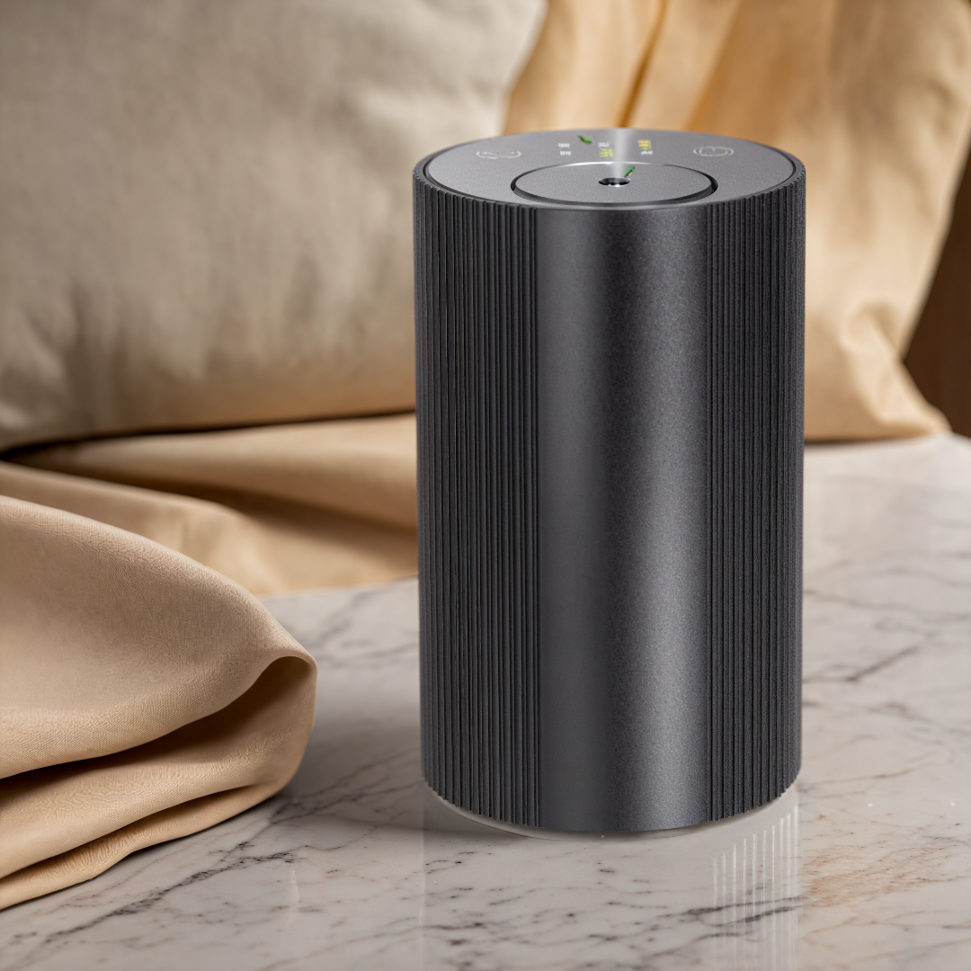 Scent Society Intro compact black diffuser with vertical ribbed texture, flat circular top, and LED indicator lights, shown standing upright on a marble surface with soft gold satin fabric and beige pillows in the background for an upscale, cozy ambiance.