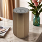 Scent Society Intro compact gold diffuser with vertical ribbed texture, flat circular top, and LED indicator lights, shown standing upright on a marble surface beside a magazine and a glass vase with pink roses, in a softly lit room with beige curtains.