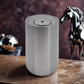Scent Society Intro compact silver diffuser with vertical ribbed texture, flat circular top, and LED indicator lights, shown standing upright on a polished brown marble surgace next to a glossy black horse sculpture and metallic decor against a deep maube background.