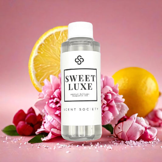 Scent Society SWEET LUXE premium diffuser fragrance oil in a clear 100ml plastic bottle with white label and cap, surrounded by pink flowers, whole lemons, red berries and scattered sugar crystals, set against a soft pink background.