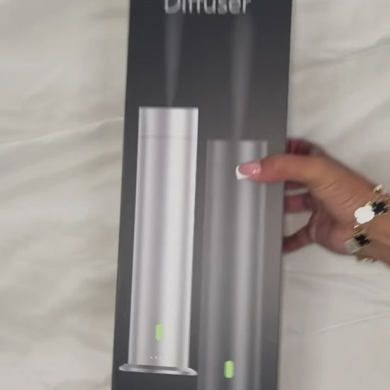 A woman’s hand opens a white box revealing a tower diffuser, user manual, remote control, power cord, and accessories neatly arranged inside. She lifts the diffuser to open it, displaying the internal bottle pre-filled with fragrance oil. She holds up the remote control, then shows the diffuser plugged in with a glowing green LED light indicating the machine is powered on and in use.