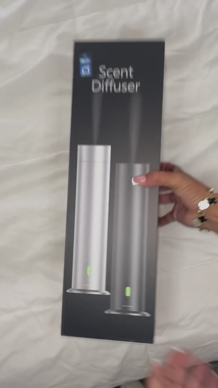 A woman’s hand opens a white box revealing a tower diffuser, user manual, remote control, power cord, and accessories neatly arranged inside. She lifts the diffuser to open it, displaying the internal bottle pre-filled with fragrance oil. She holds up the remote control, then shows the diffuser plugged in with a glowing green LED light indicating the machine is powered on and in use.